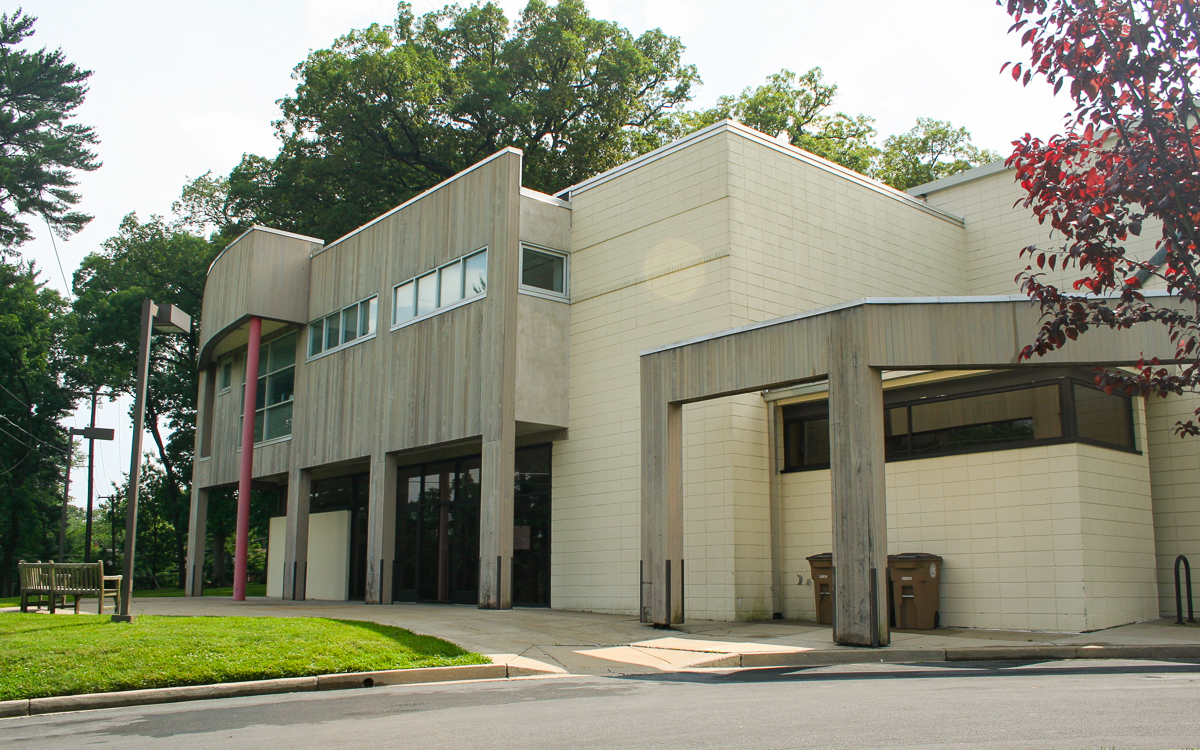 Modern two-story building with light-colored exterior, large windows, and a flat roof, surrounded by trees and a grassy area.