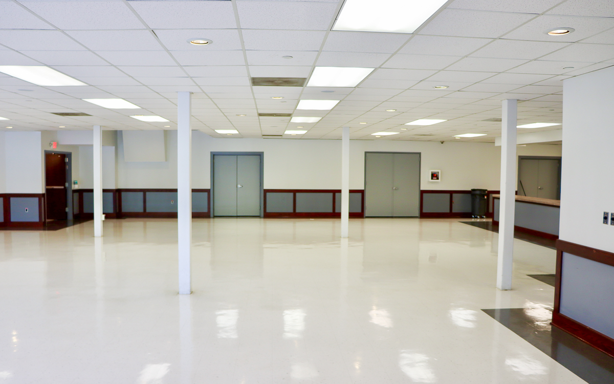 Spacious, empty room with white floors, white ceiling tiles, fluorescent lighting, columns, gray doors, and a dark wood-trimmed counter.