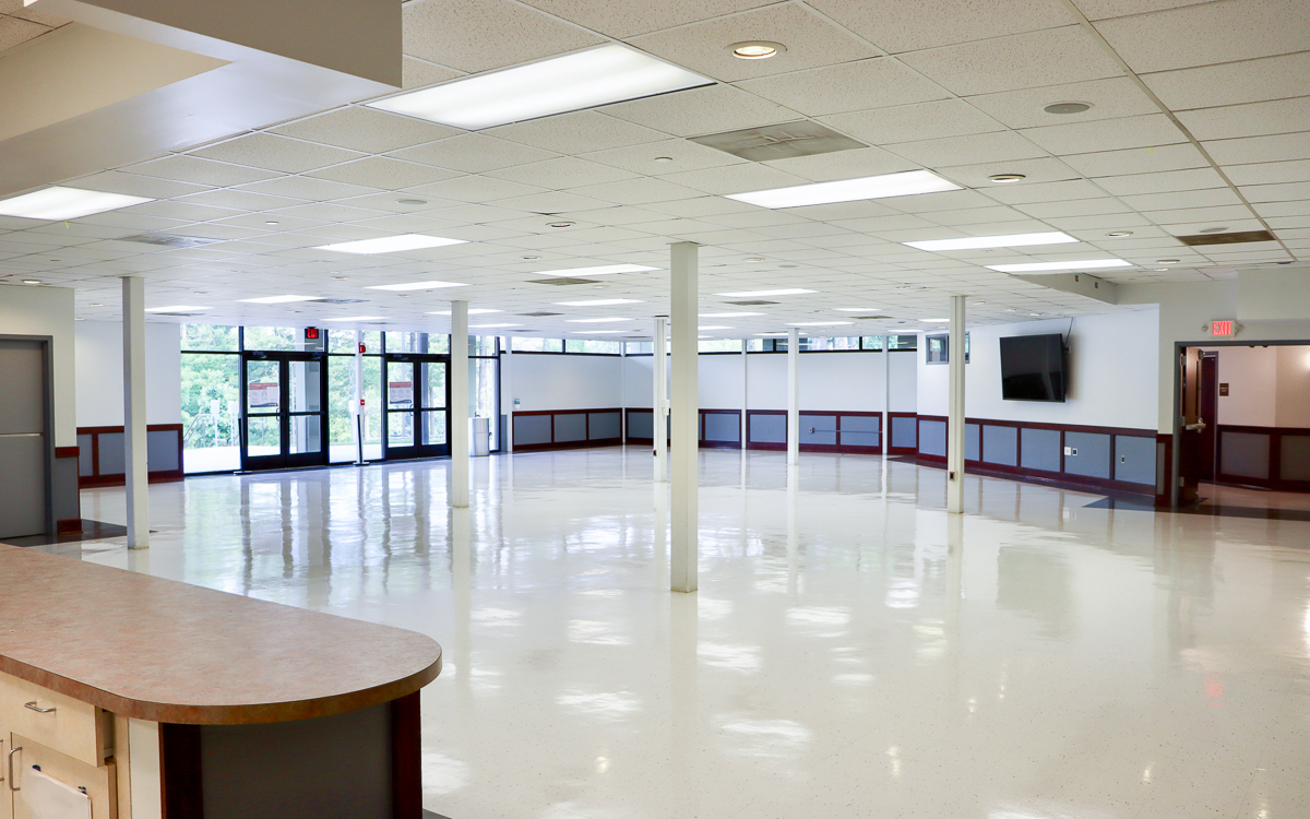Spacious, empty room with white floors, ceiling tiles, support columns, large windows, double doors, wall-mounted TV, and fluorescent lighting.