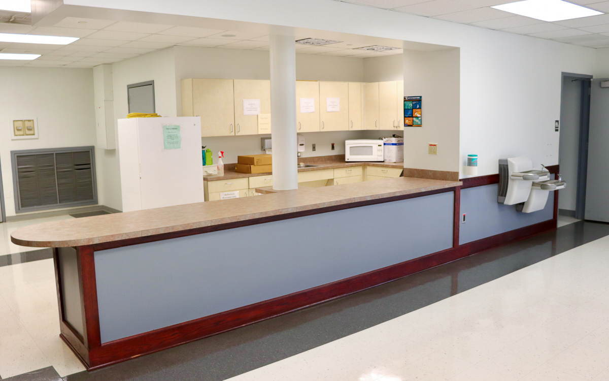 A reception counter in a clean, empty office or medical facility, with a kitchenette in the background and a water fountain on the right side.