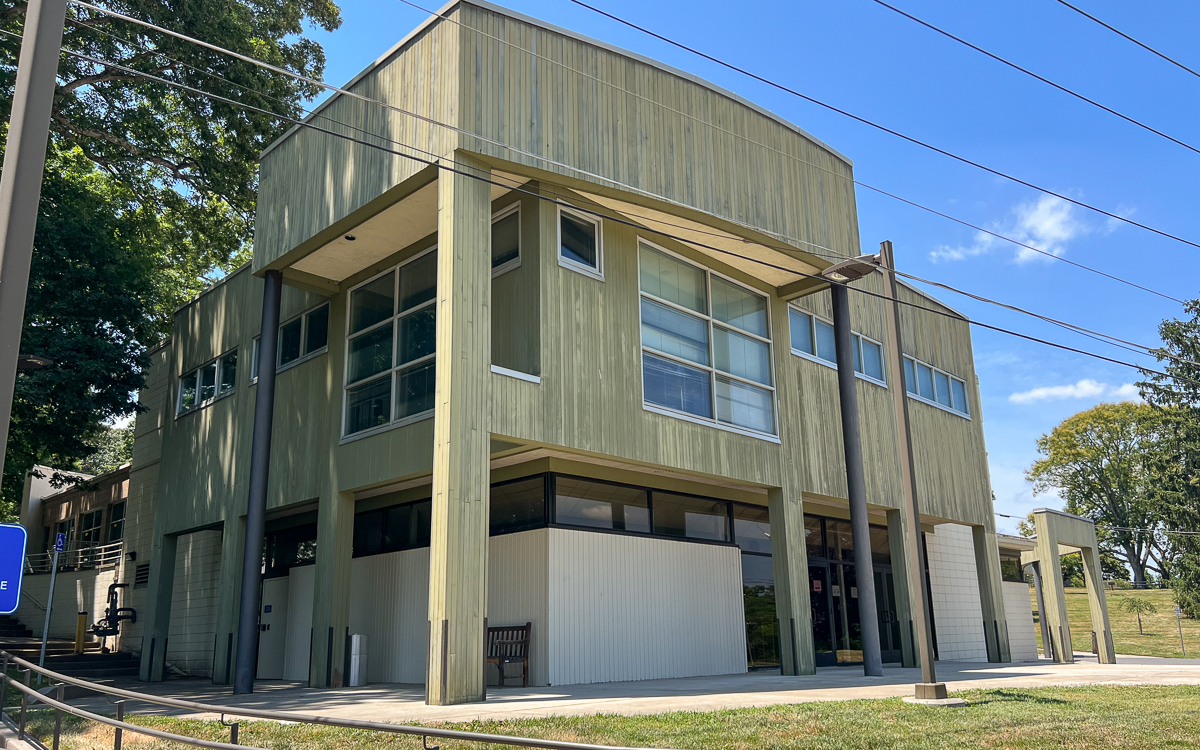 A modern two-story building with light green vertical siding, large windows, and a flat roof, surrounded by grass and trees under a clear blue sky.