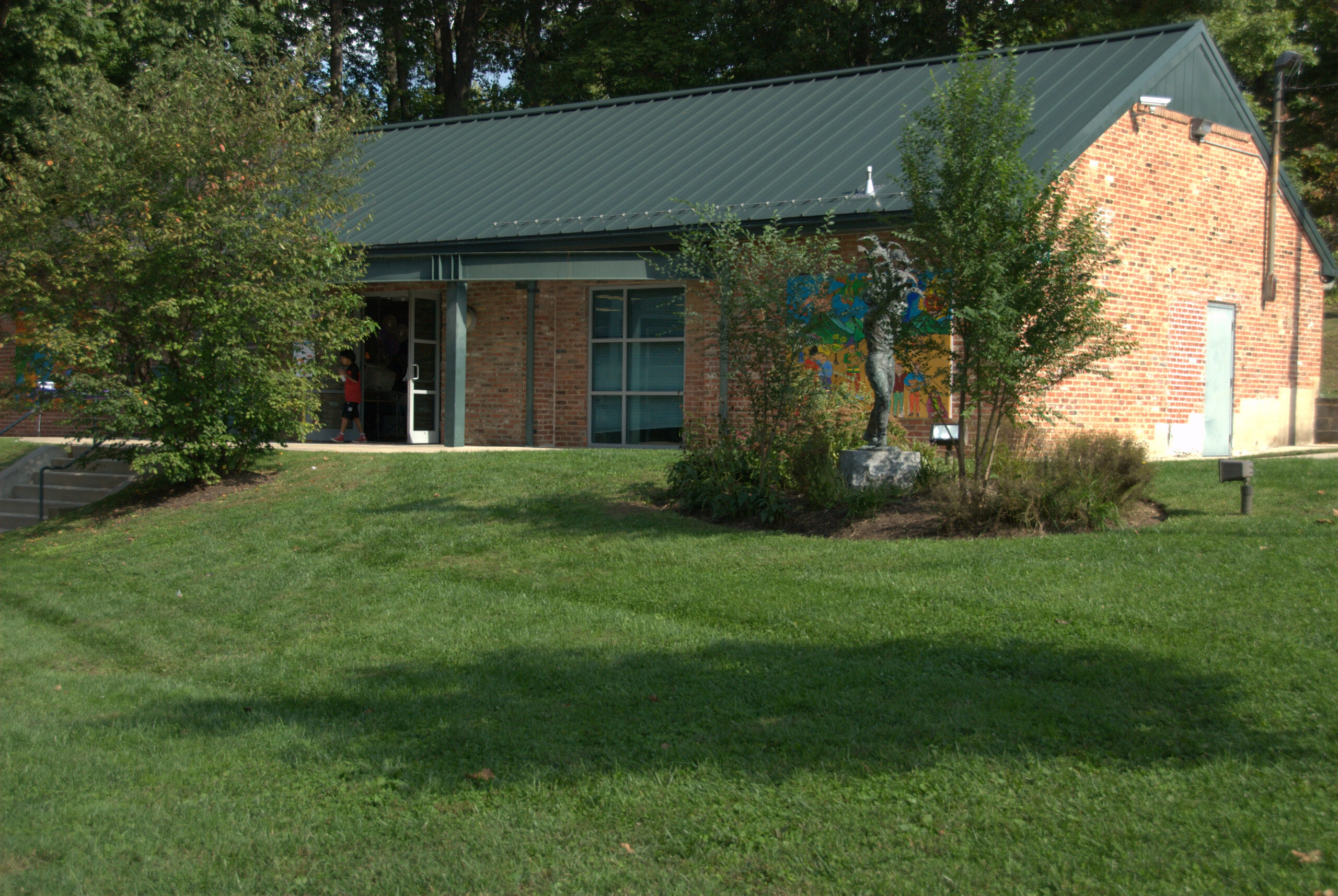 A brick building with a green metal roof, surrounded by grass and trees, has a mural on one wall and an open door with a person visible inside.
