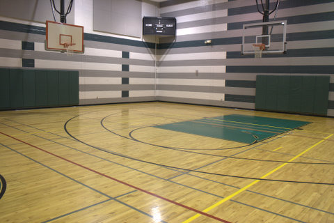Empty indoor basketball court with wood flooring, striped walls, two basketball hoops, and a scoreboard in the corner.