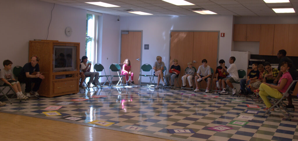 A group of children and an adult sit in a circle of chairs in a classroom with number cards arranged on the floor.