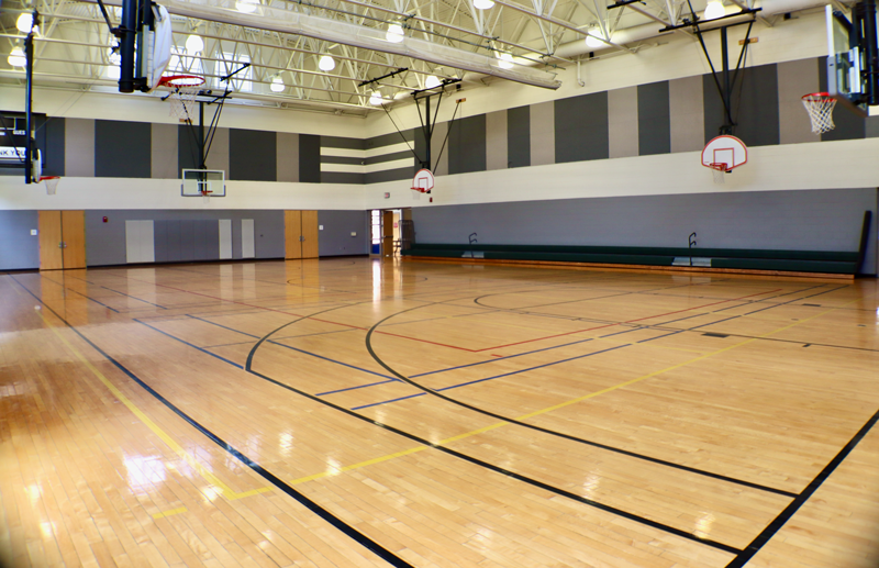 Empty indoor basketball gym with polished wooden floor, multiple hoops, and bleachers along one wall under bright overhead lighting.