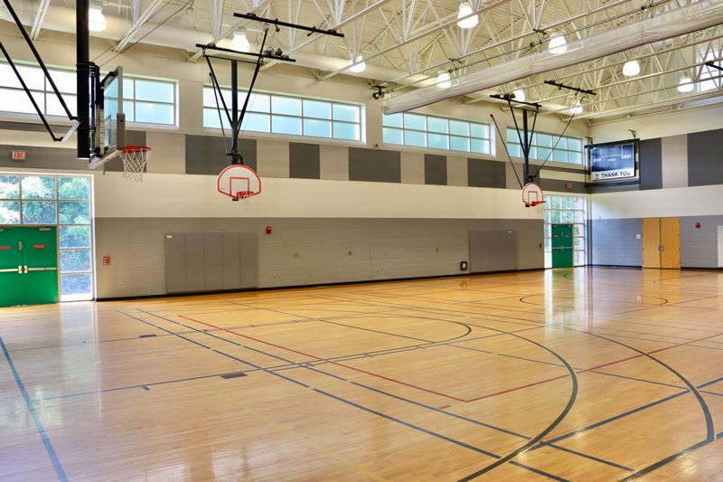 Empty indoor gymnasium with wooden floors, basketball hoops, and natural light coming through high windows. A scoreboard is mounted on the wall in the background.