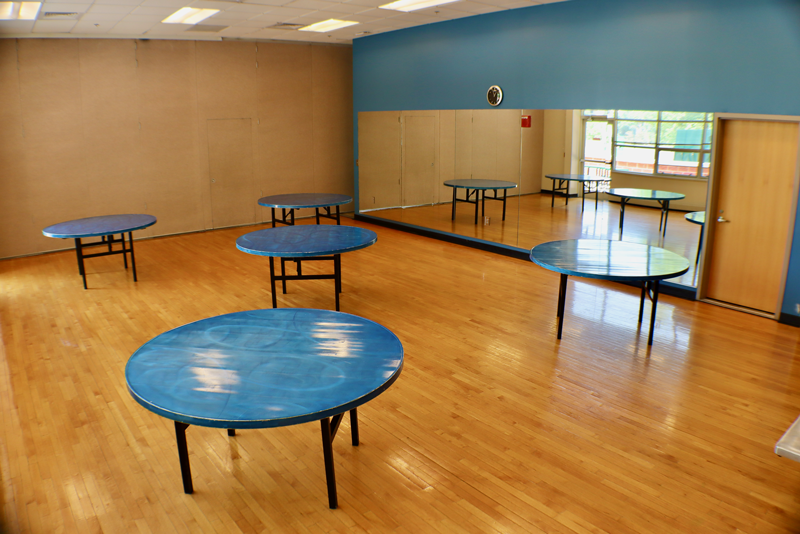 A room with five round blue tables on a wooden floor, blue walls, a large wall mirror, a clock, and windows letting in natural light.