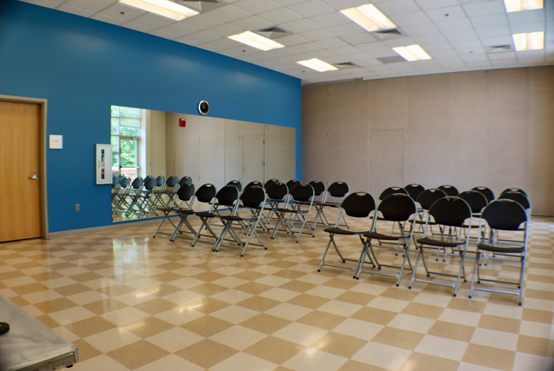 A classroom with rows of empty black folding chairs facing a wall mirror, blue accent wall, and checkered tile floor.