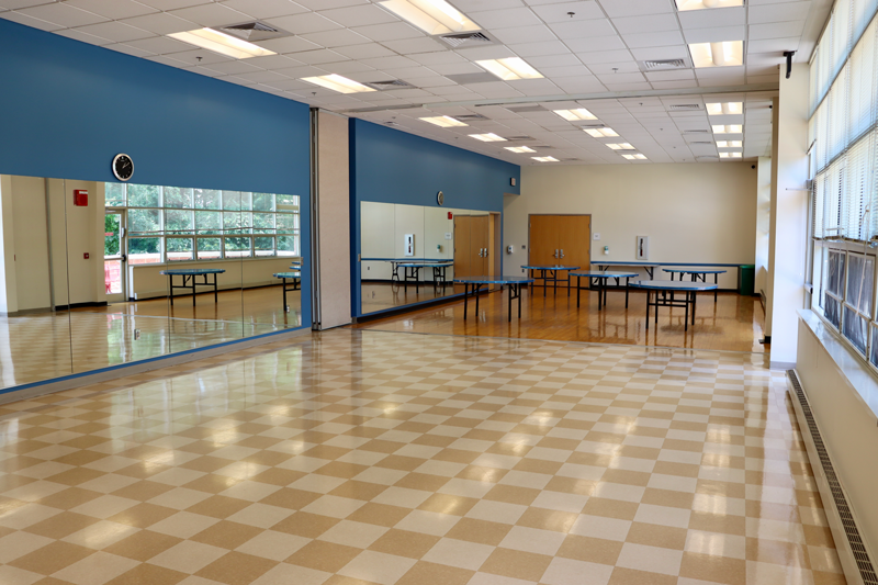 Spacious room with checkered tile floor, large wall mirrors, folding tables along the walls, fluorescent lighting, and windows on one side.