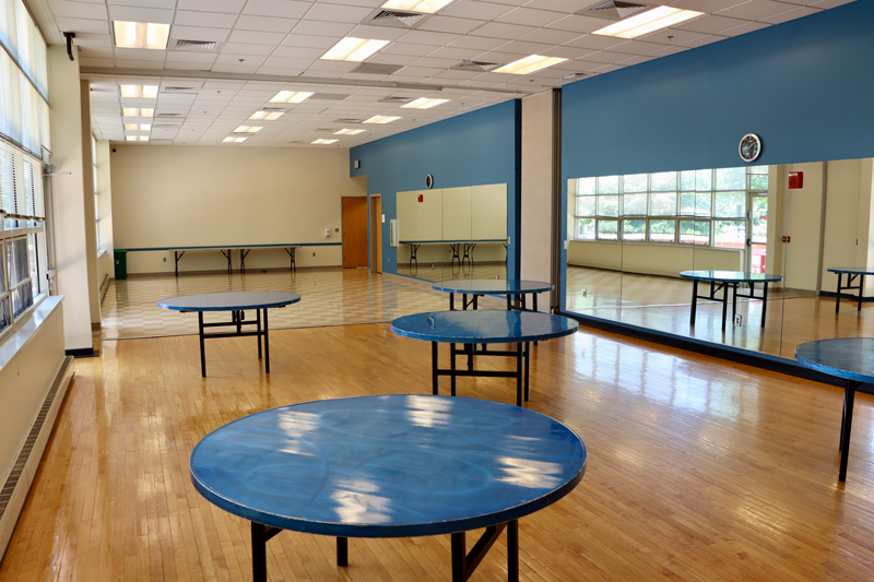 A spacious, empty room with round blue tables, wood flooring, large mirrors on one wall, and windows letting in natural light.