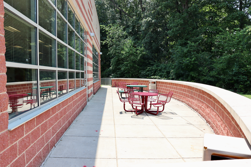 Outdoor patio with red metal tables and chairs next to a brick building; large windows on the left, trees in the background.