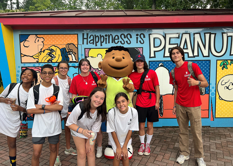 A group of kids and a mascot pose in front of a “Happiness is Peanuts” wall at an amusement park, with several kids wearing white shirts and a few in red.