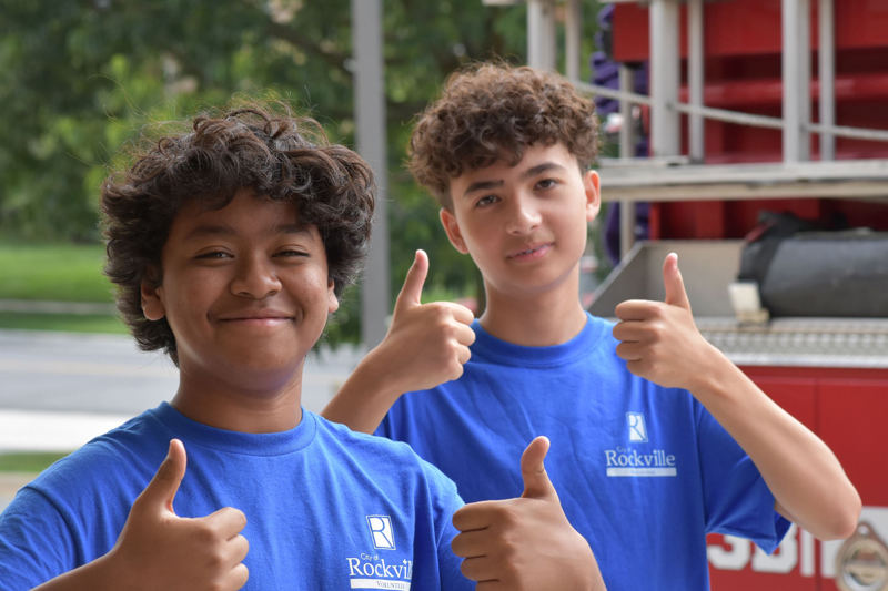 Two teens wearing blue Rockville shirts stand outside, smiling and giving thumbs up. Trees and part of a red vehicle are visible in the background.