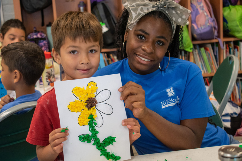 A young boy holds up a flower craft while a woman in a blue shirt sits beside him, both smiling at the camera in a classroom setting.