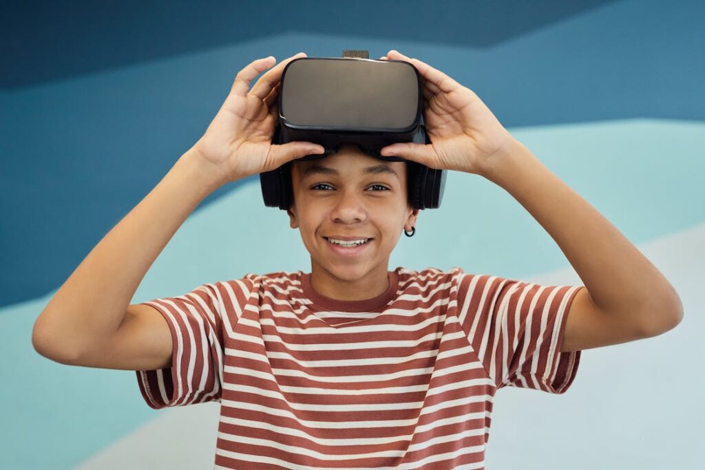 A smiling young person in a striped shirt holds a virtual reality headset on their head against a blue geometric background.