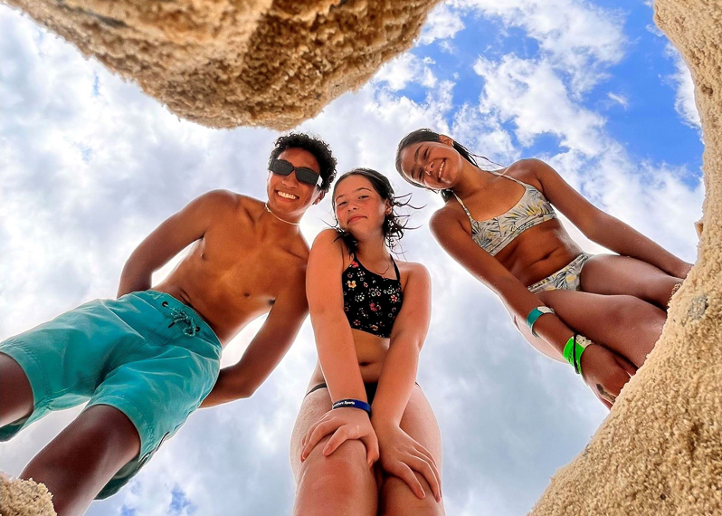 Three people in swimwear stand on sandy ground, looking down into a hole, with blue sky and clouds above them.