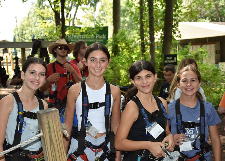 Four girls wearing harnesses and helmets stand together outdoors at an adventure park, smiling at the camera with trees and other people in the background.