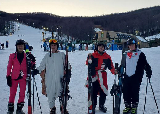 Four people in costumes and ski gear stand on a snowy ski slope at dusk, holding skis and poles, with a ski lodge and other skiers in the background.