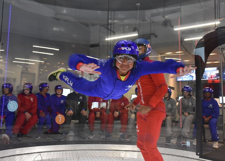 A person in blue flight gear is indoor skydiving in a wind tunnel, assisted by an instructor in red, while a group watches from the background.