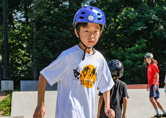 A young boy wearing a blue helmet and white skate camp T-shirt stands in a skatepark, with other children and trees in the background.