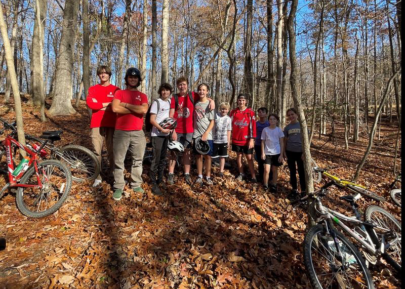 A group of people stand together in a wooded area covered with dry leaves, with several bicycles resting on the ground nearby.