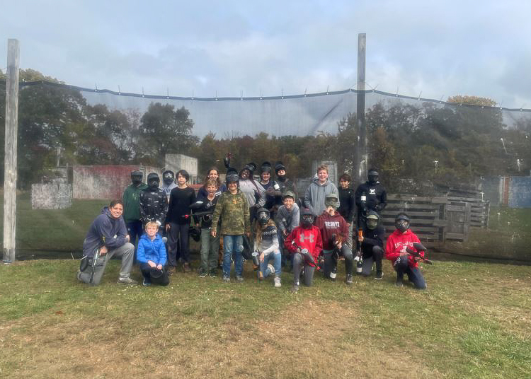 A group of people, some wearing paintball gear and masks, pose for a photo outdoors in front of a netted fence on a paintball field.