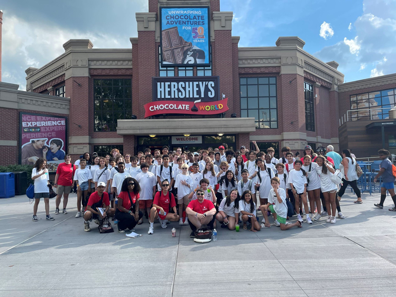 A large group of people, mostly wearing white shirts, pose for a photo outside the entrance of Hershey's Chocolate World on a sunny day.