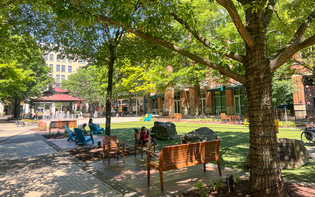 People sit on benches and chairs in a shaded urban park with trees, artificial grass, and a gazebo on a sunny day.