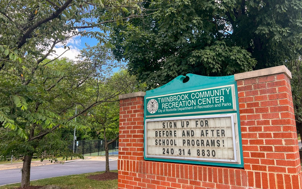 A brick sign for Twinbrook Community Recreation Center displays information about signing up for before and after school programs, with a phone number listed below. Trees and a street are in the background.