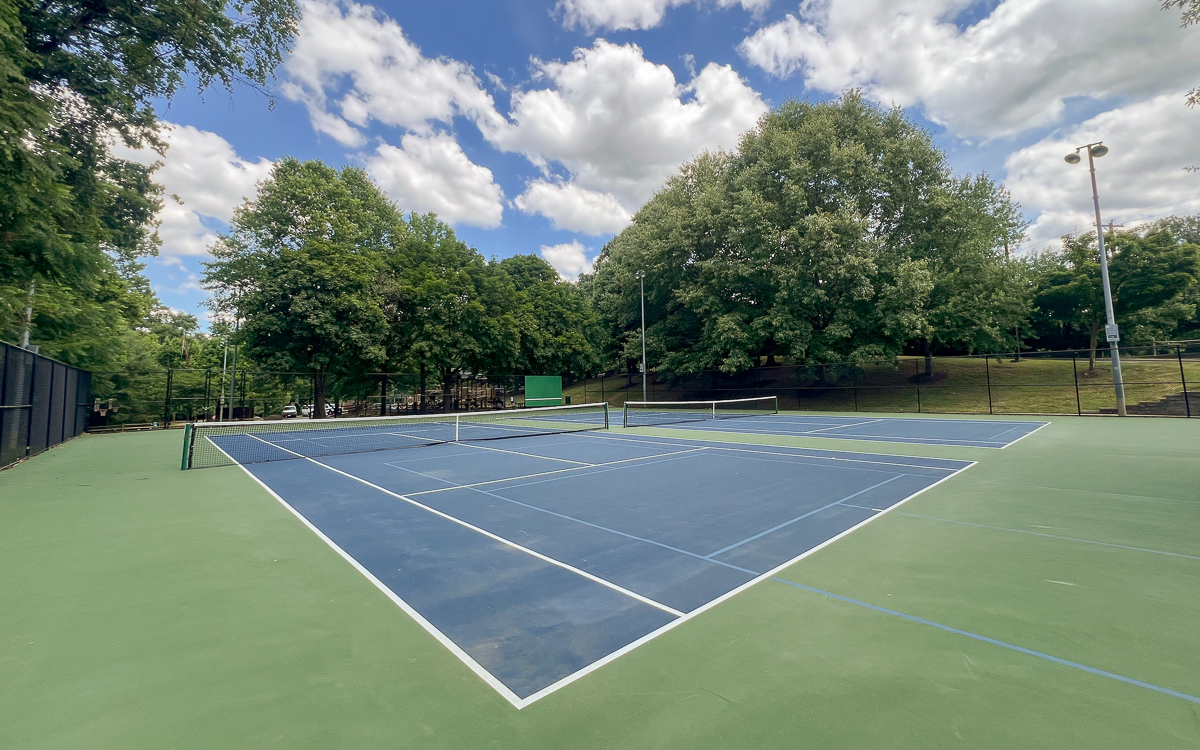 Two empty outdoor tennis courts with blue and green surfaces, surrounded by trees under a partly cloudy sky.