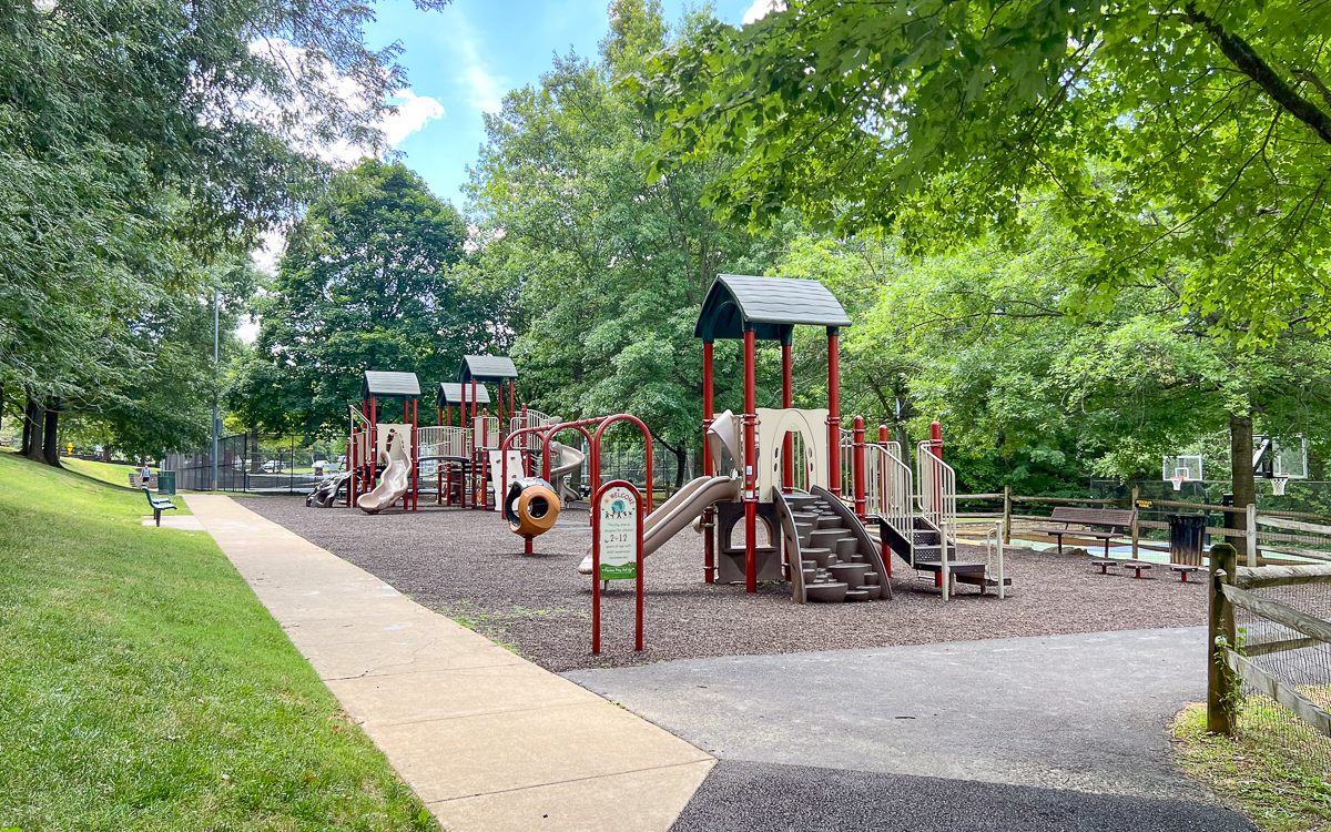 A playground with slides, climbing structures, and benches, surrounded by trees and a fence, on a sunny day.