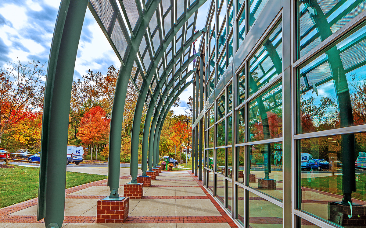 Curved metal and glass canopy outside a building, reflecting trees with autumn foliage, cars, and a person sitting on a bench.