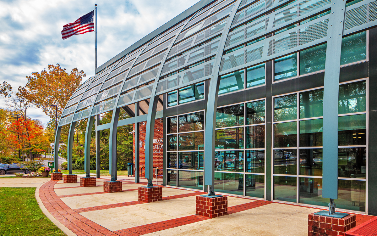 Modern building with large glass windows and metal beams, an American flag on a pole, and trees with autumn foliage in the background.