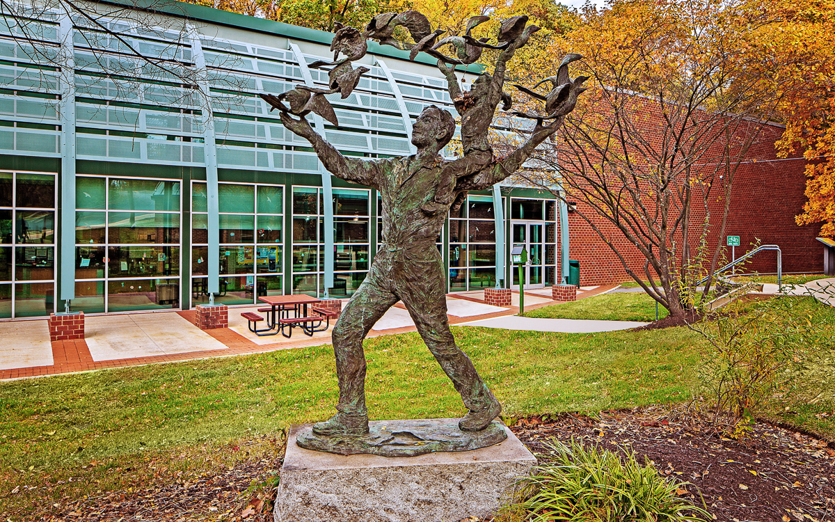 A bronze statue of a person releasing multiple birds stands on a stone base in front of a modern glass building with a brick exterior and outdoor seating.