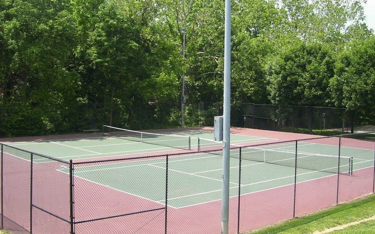 Two empty outdoor tennis courts surrounded by a chain-link fence, with trees and greenery in the background and a tall light post in the foreground.