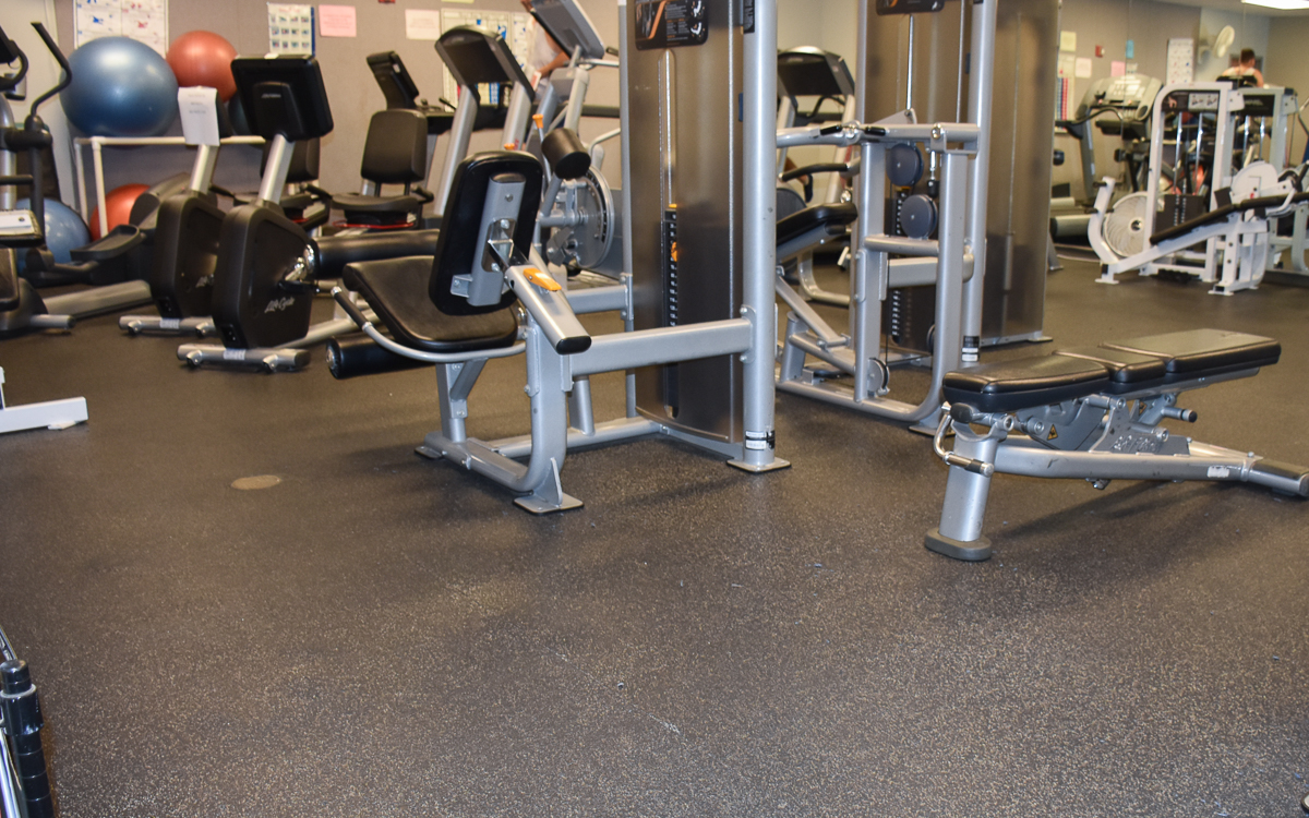 A gym with various exercise machines and equipment, including weight benches, seated row machines, and stability balls, arranged on a rubber floor.