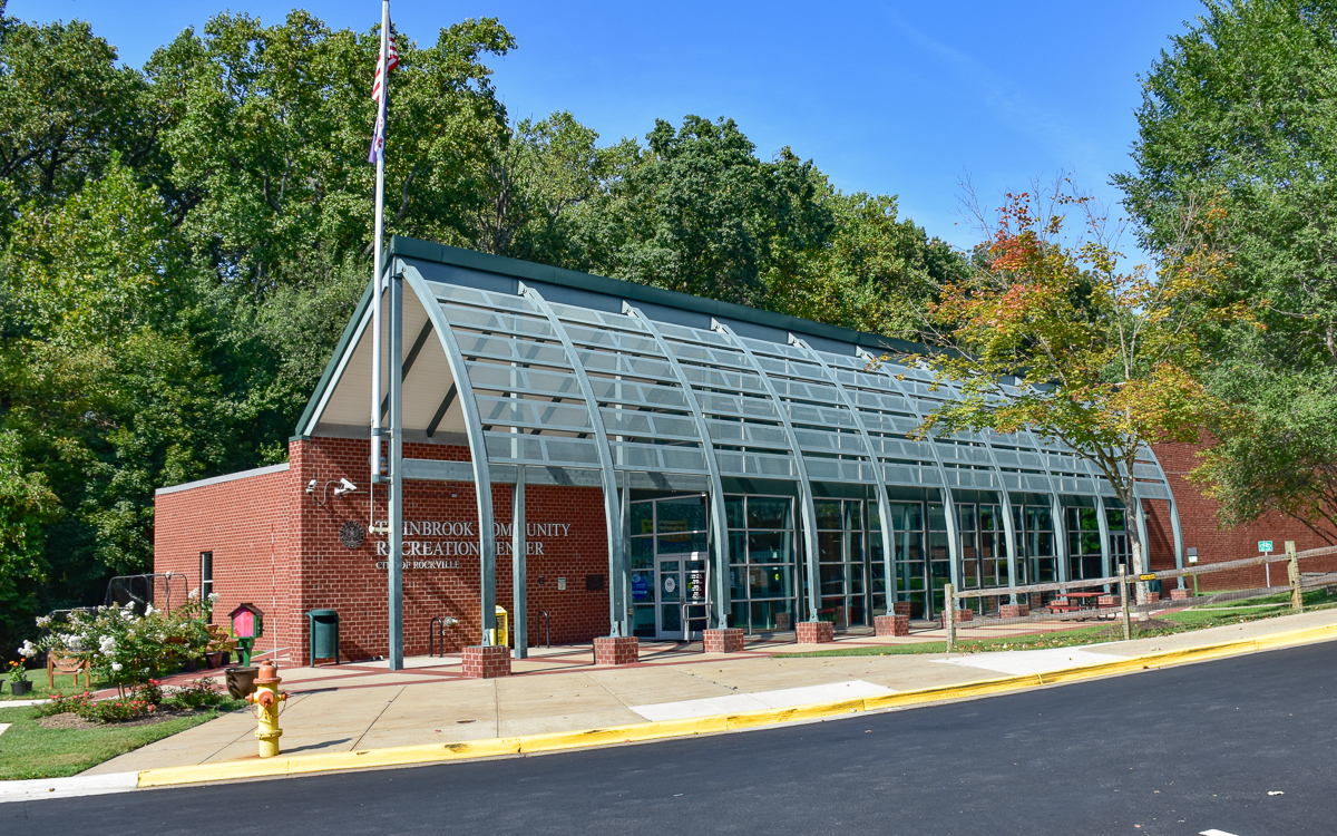 A brick and glass community recreation center building with a sloped roof, surrounded by trees and a landscaped sidewalk.