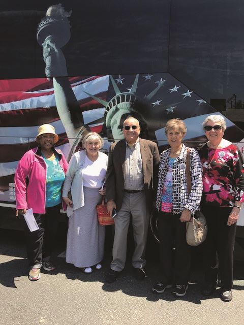 Five older adults stand together smiling in front of a bus decorated with an image of the Statue of Liberty and an American flag.