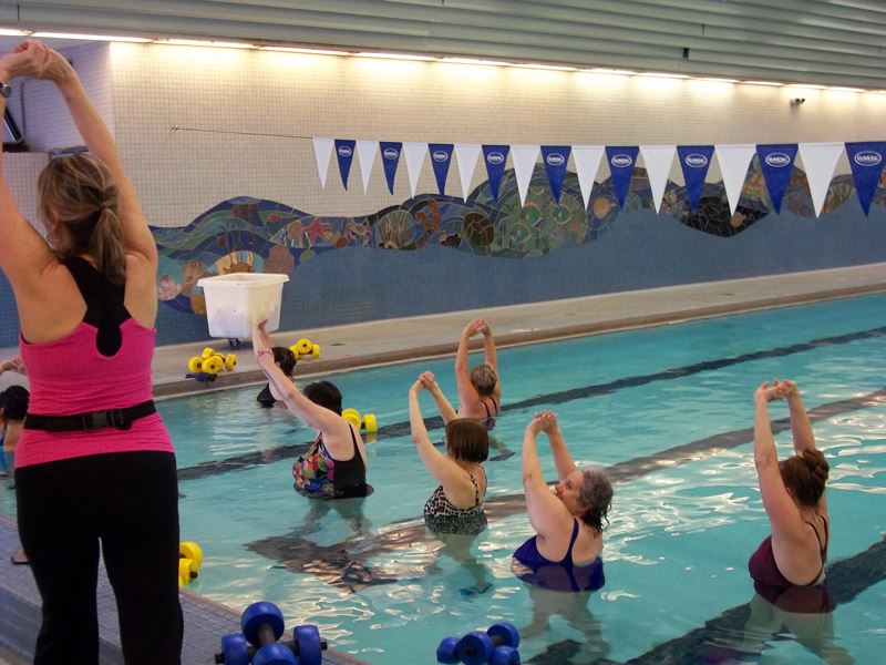 A group of adults in a swimming pool perform stretching exercises led by an instructor at the poolside.
