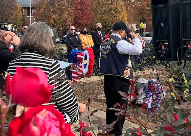 A group of people, including veterans and police officers, attend an outdoor memorial ceremony with wreaths and red flowers in the foreground.
