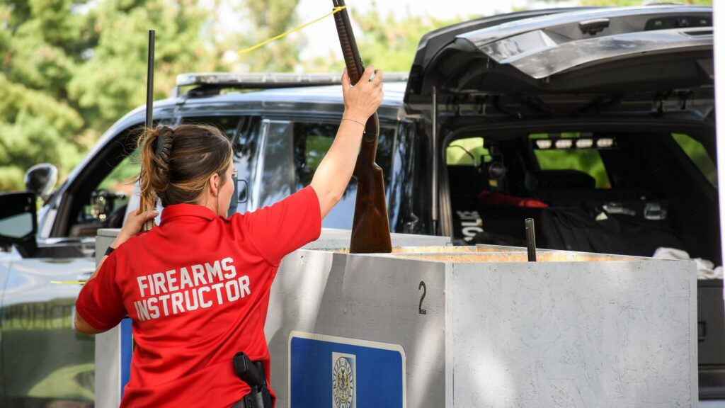 A person wearing a "Firearms Instructor" shirt handles a rifle near a drop-off bin beside an open vehicle outdoors.