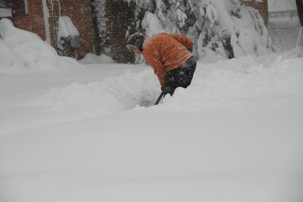 A person in an orange jacket is shoveling snow during heavy snowfall, surrounded by deep snowdrifts near a brick building and trees.