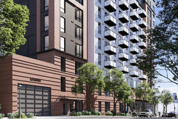 A modern multi-story apartment building with balconies, large windows, and a brick facade, surrounded by trees and a sidewalk on a sunny day.