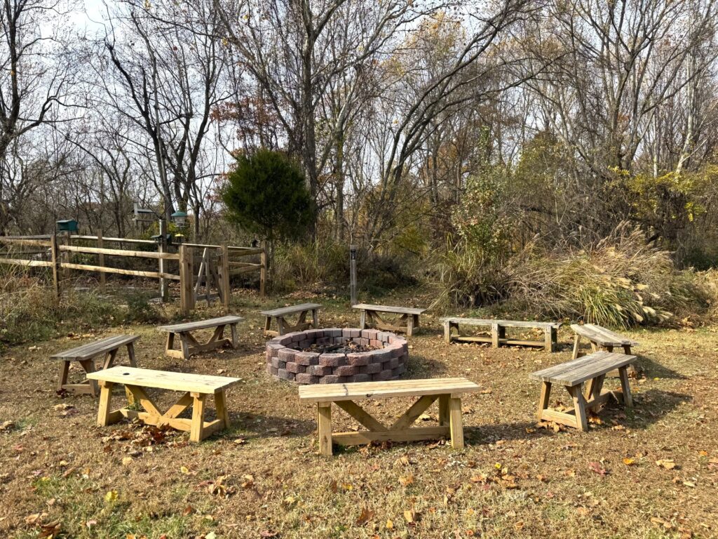 Six wooden benches arranged in a circle around a brick fire pit in a grassy clearing, with trees and a wooden fence in the background.