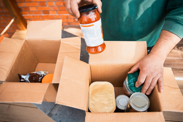 Person packing food items, including canned goods and a jar of sauce, into cardboard boxes on a table.