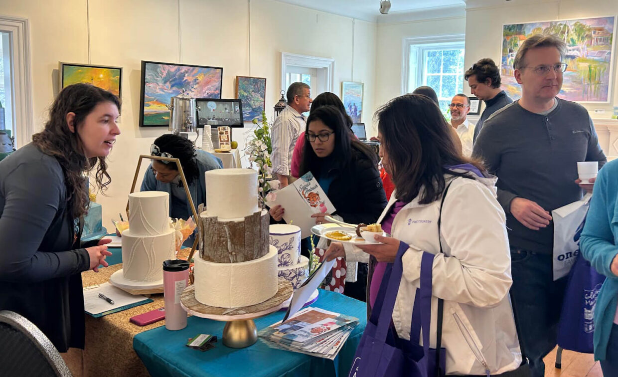 People gather around a decorated table with cakes and brochures at an indoor event, with paintings displayed on the walls in the background.
