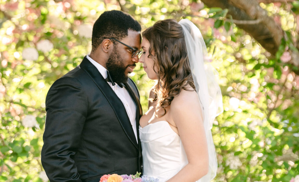 A bride in a white dress and veil holds a bouquet while standing closely with a groom in a black tuxedo, facing each other outdoors among greenery and flowers.
