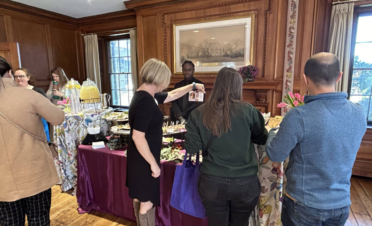 Several people gather around a buffet table with assorted food and drinks in a wood-paneled room with large windows and floral decorations.