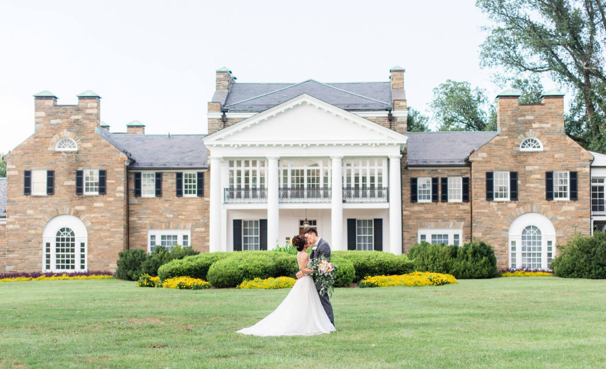 A bride and groom embrace in front of a large, historic mansion with stone walls, white columns, and manicured gardens.