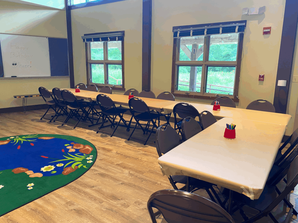 A classroom with two long tables and chairs, a colorful rug, art supplies on tables, windows letting in natural light, and a whiteboard with a birthday message.
